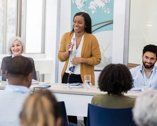 A facilitator stands to lead a live training session, microphone in hand, while participants follow along with printed materials at the table